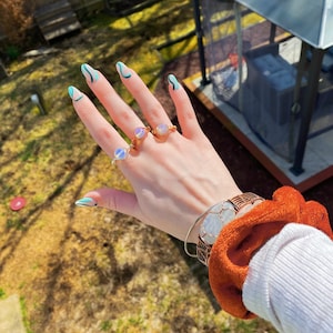 May include: A hand with blue and white nail polish is wearing three rings with iridescent stones. The hand is also wearing a bracelet with a large, white, iridescent stone.