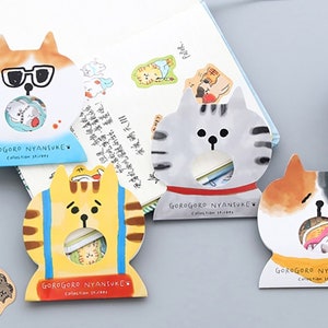 Könnte beinhalten: Vier farbenfrohe Aufkleber-Pakete in Katzenform mit einem Loch in der Mitte, um die Aufkleber im Inneren zu zeigen. Jedes Paket hat ein anderes Katzendesign und den Text "GOROGORO NYANSUKE Collection Stickers" unten.