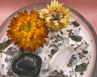 May include: A white candle with dried flowers, green leaves, and clear and dark stones embedded in the wax. The candle is in a clear glass jar.
