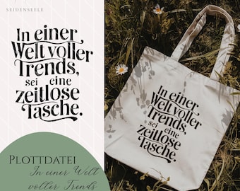 Könnte beinhalten: Eine weiße Stofftasche mit schwarzem Text, der "In einer Welt voller Trends, sei eine zeitlose Tasche." lautet. Die Tasche liegt auf einem Bett aus grünem Gras und weißen Blumen.
