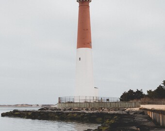 May include: A red and white lighthouse stands tall against a gray sky. The lighthouse is surrounded by rocks and water.