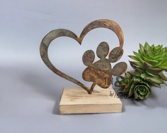 May include: A metal heart-shaped sculpture with a paw print inside. The sculpture is on a wooden base. The metal is rusted and has a brown and green patina.
