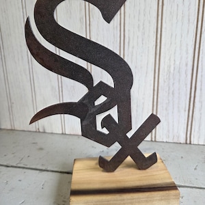 May include: A metal sculpture of the Chicago White Sox logo, featuring a stylized "S" and "X" in a dark, weathered finish. The sculpture is mounted on a rectangular wooden base, showcasing a natural wood grain.