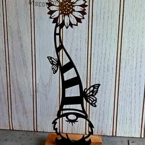 May include: A black metal garden gnome silhouette with a sunflower head and butterfly accents. The gnome's striped hat is supported by a wooden base. The image is set against a white wood-paneled background.