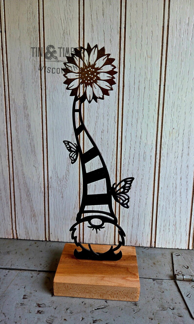 May include: A black metal garden gnome silhouette with a sunflower head and butterfly accents. The gnome's striped hat is supported by a wooden base. The image is set against a white wood-paneled background.