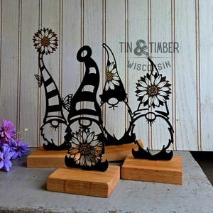 May include: Four black metal gnome figurines with sunflower and butterfly accents, mounted on wooden blocks. The gnomes have striped and solid hats. The text "Tin & Timber Wisconsin" is visible in the background.