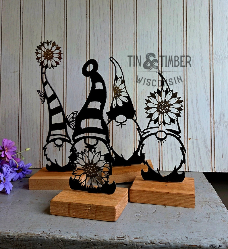 May include: Four black metal gnome figurines with sunflower and butterfly accents, mounted on wooden blocks. The gnomes have striped and solid hats. The text "Tin & Timber Wisconsin" is visible in the background.