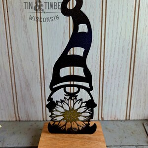 May include: A black metal gnome silhouette with a striped hat and sunflower detail, mounted on a wooden base. The image includes the text "TIN & TIMBER WISCONSIN".