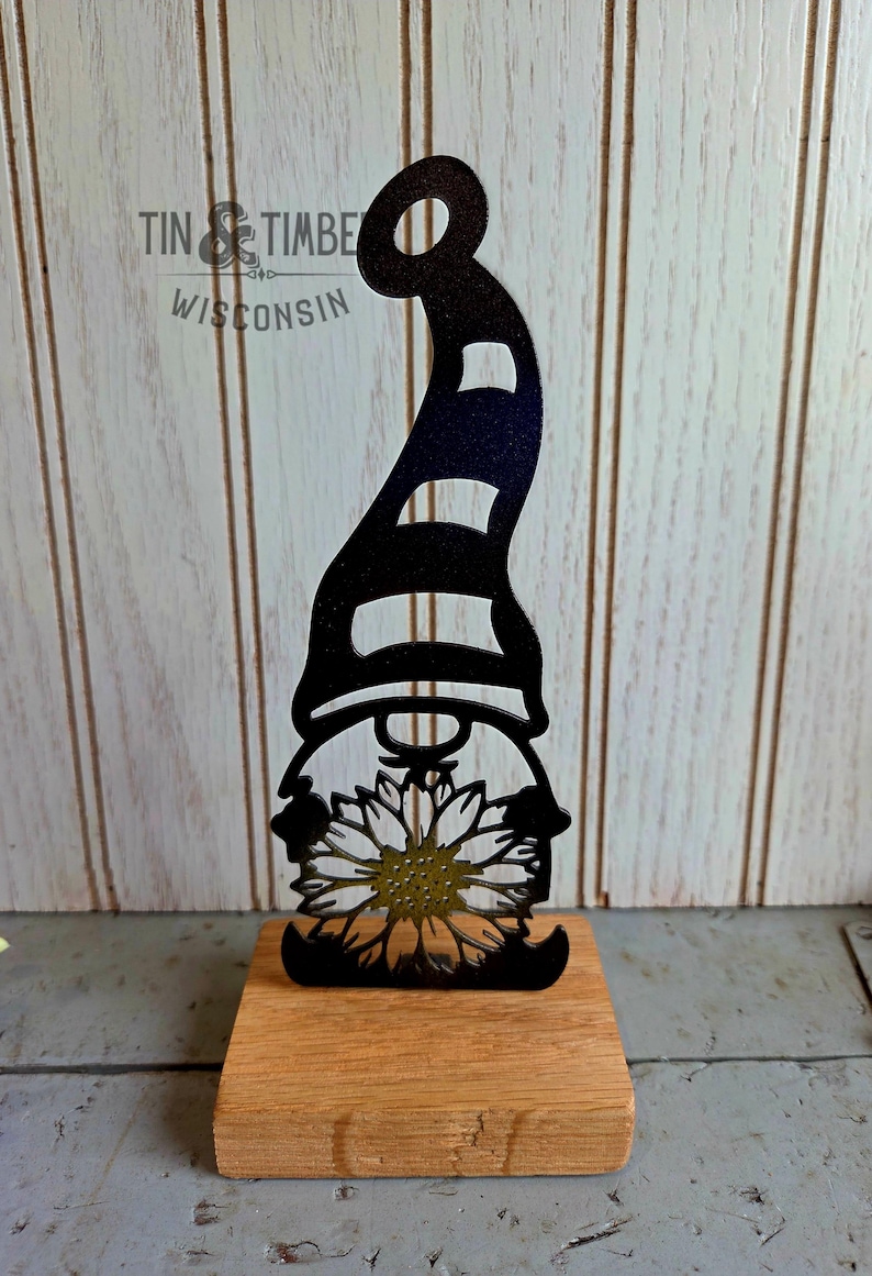 May include: A black metal gnome silhouette with a striped hat and sunflower detail, mounted on a wooden base. The image includes the text "TIN & TIMBER WISCONSIN".