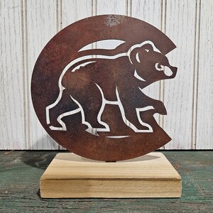Puede incluir: Una silueta de oso de metal oxidado recortada sobre una base de madera. El oso está mirando hacia la derecha y está en posición de caminar. El metal es de color marrón oscuro con un acabado rústico.