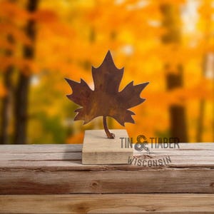 May include: A metal maple leaf sculpture with a rustic brown finish, mounted on a small wooden block. The sculpture is set against a blurred background of fall foliage. The text "TIN & TIMBER WISCONSIN" is visible.