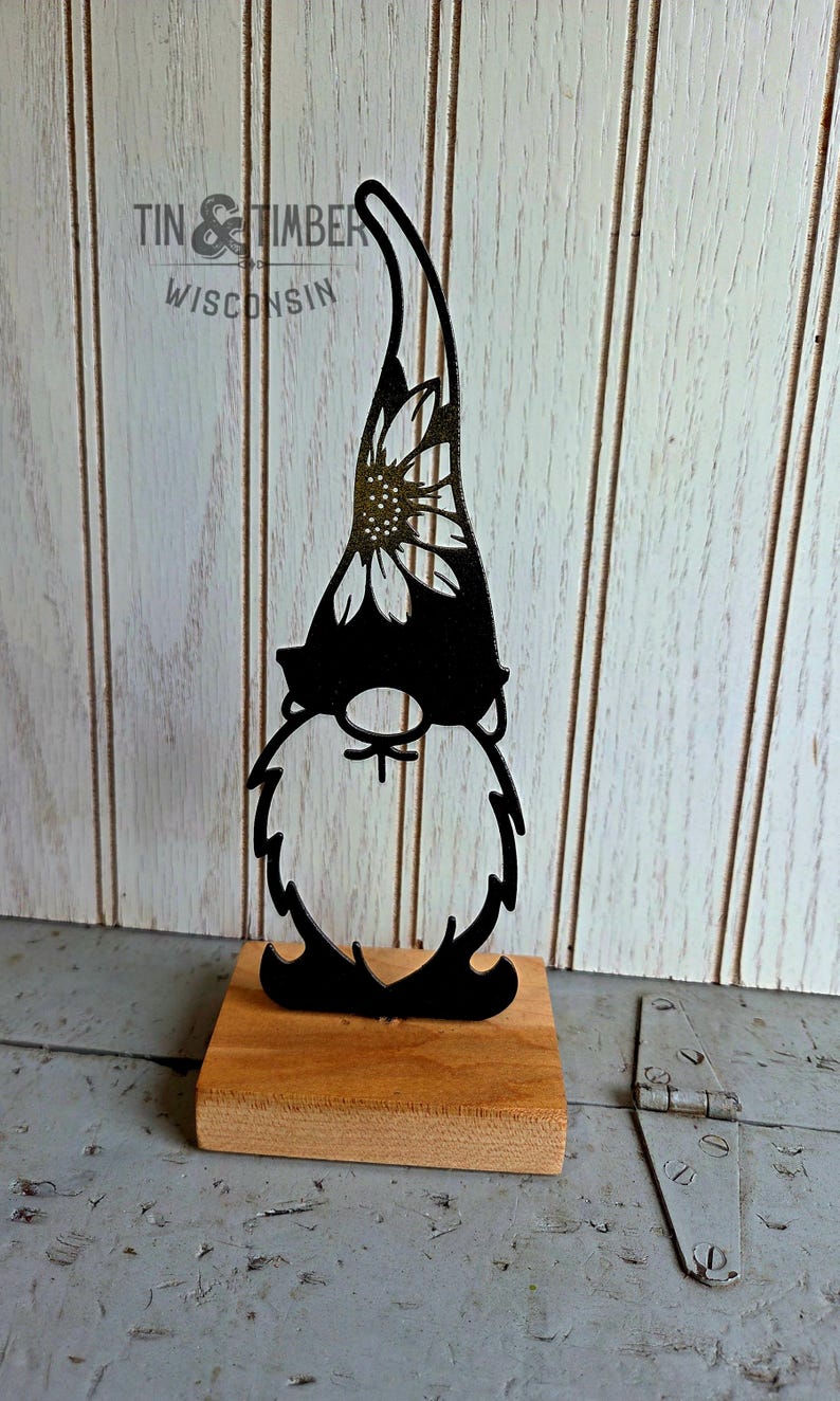 May include: Black metal gnome silhouette with a sunflower design on the hat, mounted on a small wooden base. The gnome has a long beard and a pointed hat. The text "TIN & TIMBER WISCONSIN" is visible in the background.