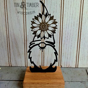 May include: Black metal gnome figurine holding a sunflower, mounted on a wooden base. The gnome has a long beard and a pointed hat. The sunflower has detailed petals and a textured center. The words "TIN & TIMBER WISCONSIN" are in the background.