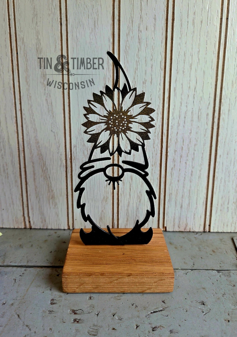 May include: Black metal gnome figurine holding a sunflower, mounted on a wooden base. The gnome has a long beard and a pointed hat. The sunflower has detailed petals and a textured center. The words "TIN & TIMBER WISCONSIN" are in the background.