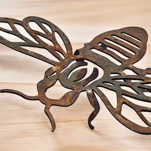 Può includere: Una scultura di ape in metallo arrugginito con intricati tagli. L'ape è in una posa di volo con le ali spiegate. La scultura ha un aspetto rustico e invecchiato.