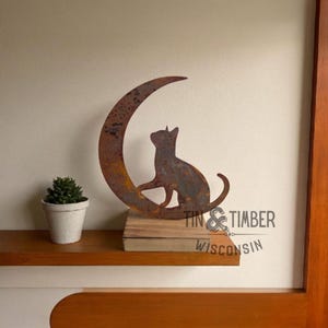 May include: A rustic metal cat silhouette sits atop a wooden base, perched on a crescent moon. The cat and moon have a weathered, brown finish. The words "TIN & TIMBER WISCONSIN" are printed on the base.