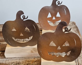 Op de afbeelding: Drie metalen jack-o'-lantern decoraties met uitgesneden ogen, neuzen en monden. Elke pompoen is gemonteerd op een lichtgekleurde houten basis. De pompoenen hebben een rustieke, bruine afwerking, perfect voor Halloween.
