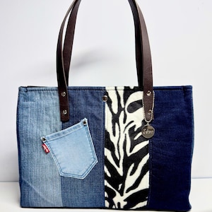 Può includere: Una borsa tote in denim con manici in pelle marrone. La borsa presenta un design patchwork con varie tonalità di denim blu, un pannello con stampa zebrata e una piccola tasca in denim con un'etichetta rossa. Un ciondolo d'argento è appeso.