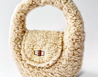 Könnte beinhalten: Eine kleine, flauschige Handtasche in Cremefarbe mit einem Henkel oben und einem goldenen Verschluss.