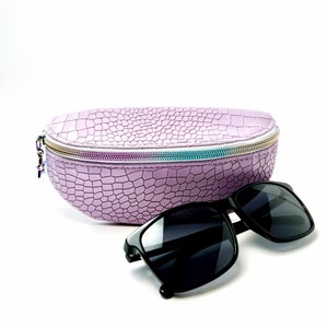 May include: A purple crocodile-embossed hard case for sunglasses with a zipper closure. The case is open and a pair of black sunglasses are lying next to it.