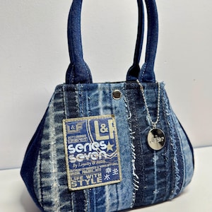Könnte beinhalten: Jeans-Handtasche mit Patchwork-Design. Die Tasche hat ein rechteckiges Etikett mit Text und einem Stern, einen silberfarbenen Anhänger und dunkelblaue Griffe. Die Abmessungen der Tasche betragen ca. 20 cm Höhe und 25 cm Breite.