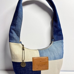 May include: A patchwork denim shoulder bag with a curved shape. The bag features a mix of light and dark blue denim panels, with a beige canvas section. A small metal tag and a leather patch with text are attached.