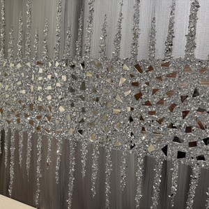 May include: A silver and grey abstract wall art piece with a textured, glittery finish. The art features a band of silver glitter and small mirrored pieces.