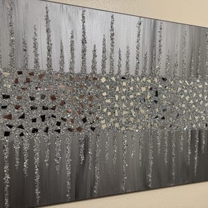 May include: A silver and grey abstract wall art piece with a textured, glittery finish. The art features a horizontal band of small, mirrored pieces, creating a shimmering effect.