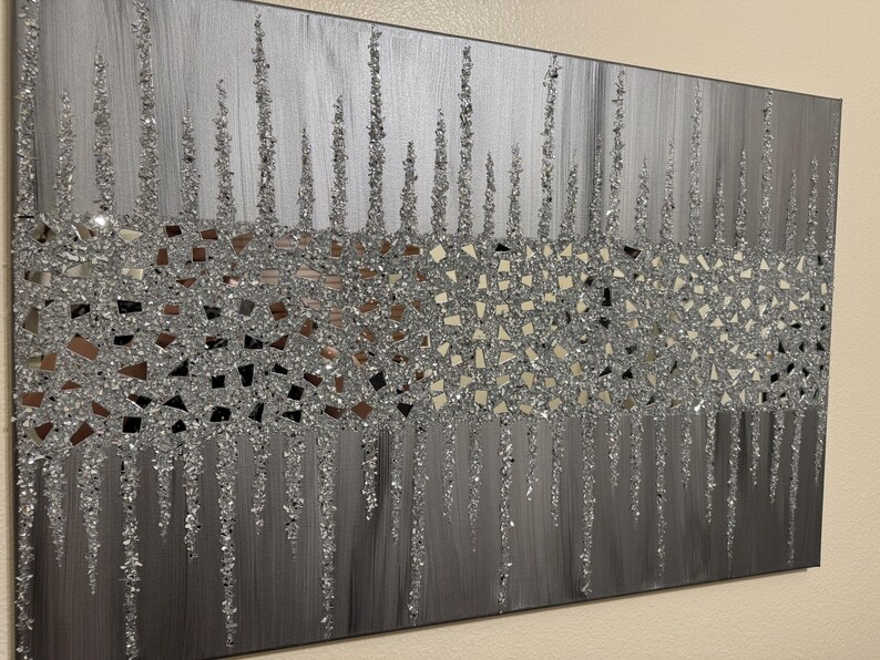 May include: A silver and grey abstract wall art piece with a textured, glittery finish. The art features a horizontal band of small, mirrored pieces, creating a shimmering effect.