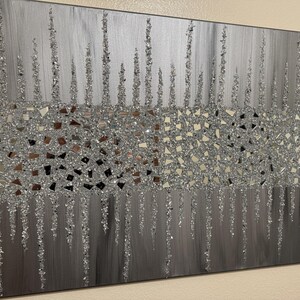 May include: A silver and grey abstract wall art piece with a textured glitter finish. The art features a design of vertical lines with a band of small, irregular silver shapes.