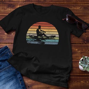 Puede incluir: Camiseta negra con un diseño retro de un remero en un bote, sobre un colorido atardecer. El diseño tiene un aspecto vintage. También se ven unas gafas de sol y una pequeña planta suculenta.