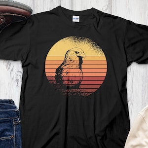 Könnte beinhalten: Schwarzes T-Shirt mit einem Retro-Grafikdruck eines braunen Adlers vor einem Sonnenuntergang. Der Adler blickt nach links und hat einen gelben Schnabel und gelbe Krallen. Der Sonnenuntergang ist ein Farbverlauf von Orange, Gelb und Rot.