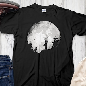 May include: Black t-shirt featuring a graphic of a person standing in front of a full moon with trees in the foreground.
