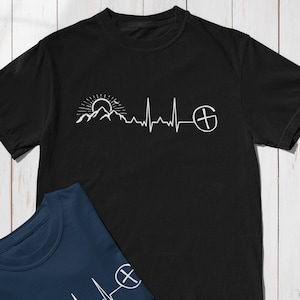 Könnte beinhalten: Schwarzes T-Shirt mit weißer Grafik eines Bergmassivs, einer Sonne und einer Herzschlaglinie, die mit einem Kreis- und Kreuzsymbol endet. Ein zweites marineblaues T-Shirt ist teilweise sichtbar.