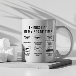 以下が含まれることがあります： 白いセラミック製のマグカップに、黒いデザインで「Things I do in my spare time」と書いてあり、ドローンに関連する活動がリストされています。マグカップは灰色のコースターの上に置かれています。
