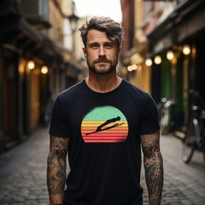 Könnte beinhalten: Ein schwarzes Kurzarm-T-Shirt mit einer Grafik eines Skispringers in Silhouette vor einem kreisförmigen Sonnenuntergangshintergrund mit horizontalen Streifen in Grün-, Gelb-, Orange- und Rottönen, mittig auf der Brust des Kleidungsstücks.
