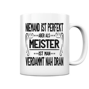 以下が含まれることがあります： 黒い文字で「NIEMAND IST PERFEKT ***ABER ALS*** MEISTER ***IST MAN*** VERDAMMT NAH DRAN」と書かれた白いセラミック製のコーヒーカップ