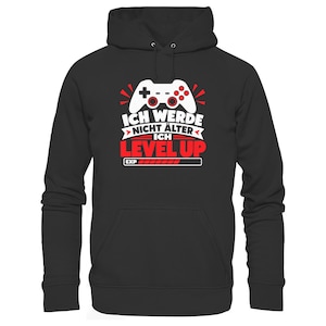 Könnte beinhalten: Ein schwarzer Kapuzenpullover mit einer weißen Grafik eines Videospiel-Controllers über dem Text "ICH WERDE NICHT ÄLTER ICH LEVEL UP" in Weiß und Rot, sowie einer mit "EXP" beschrifteten Erfahrungsleiste am unteren Rand.