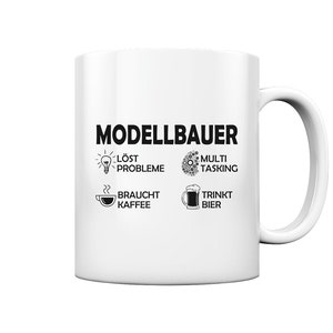 Op de afbeelding: Witte keramische mok met zwarte tekst die "Modellbauer" luidt en vier pictogrammen. De pictogrammen tonen een gloeilamp met de tekst "löst probleme", een tandwiel met de tekst "multi tasking", een koffiekopje met de tekst "braucht kaffee" en een bierglas met de tekst "trinkt bier".