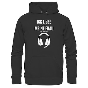 Puede incluir: Una sudadera con capucha negra que presenta texto en blanco que dice ICK LIEBE es, wenn MEINE FRAU mich Zocken lässt, acompañado de una ilustración gráfica blanca de auriculares para juegos con micrófono debajo del texto.