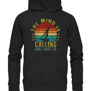 Può includere: Felpa con cappuccio nera con una grafica vintage di una barca a vela e il testo "The Wind Is Calling And I Must Go".
