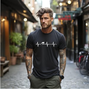 Könnte beinhalten: Ein schwarzes Kurzarm-T-Shirt mit einem weißen Grafikdruck einer Herzschlaglinie, oder EKG, bei dem eine kleine Silhouette einer Person beim Skispringen in die Mitte des Wellenmusters im Brustbereich integriert ist.