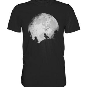 Może przedstawiać: Czarny t-shirt z grafiką psa stojącego przed dużym, białym księżycem o fakturowanej powierzchni. Księżyc jest otoczony drzewami.