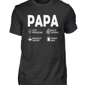 Può includere: Maglietta nera con testo bianco che dice "Papa" e quattro icone con testo. Le icone rappresentano una lampadina con il testo "löst probleme", una tazza di caffè con il testo "braucht kaffee", un ingranaggio con il testo "multi tasking", e una boccale di birra con il testo "trinkt bier".