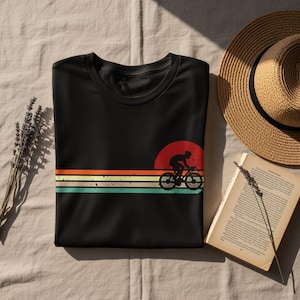 Könnte beinhalten: Schwarzes T-Shirt mit einer Radfahrer-Silhouette und einem roten Sonnendesign. Das Design umfasst orange, gelbe und türkisfarbene horizontale Streifen. Ein Strohhut und ein offenes Buch sind im Hintergrund zu sehen.