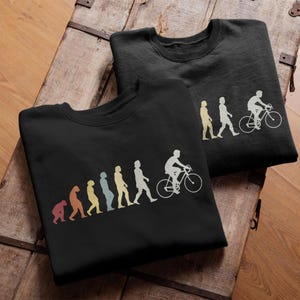 Può includere: Due felpe nere con una grafica evolutiva in stile retrò. La grafica raffigura l'evoluzione dell'uomo, culminando con una persona in bicicletta. Il design utilizza tonalità di rosso, arancione, giallo, blu e bianco.