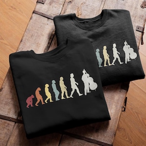 Może przedstawiać: Dwie czarne bluzy z grafiką ewolucji w stylu retro. Grafika przedstawia progresję od małpy do osoby grającej na kontrabasie, w kolorowych sylwetkach. Bluzy są prezentowane na drewnianej powierzchni.