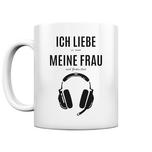 Könnte beinhalten: Weiße Keramiktasse mit schwarzem Text, der "Ich liebe (Herzsymbol) meine Frau, mich zocken lässt" lautet. Der Text ist in einer verspielten Schriftart. Unter dem Text befindet sich eine Schwarzweißillustration eines Gaming-Headsets mit Mikrofon.