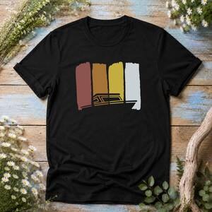 Könnte beinhalten: Ein schwarzes T-Shirt mit kurzen Ärmeln und einem Grafikdruck auf der Brust. Das Design besteht aus vier vertikalen, rechteckigen Pinselstrichen in den Farben Kastanienbraun, Braun, Gelb und Weiß sowie einer einfachen Boots-Strichzeichnung.