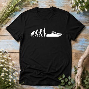Könnte beinhalten: Ein schwarzes T-Shirt mit kurzen Ärmeln und einem weißen Grafikdruck auf der Brust. Das Design zeigt die evolutionäre Entwicklung von Primaten hin zu einer menschlichen Figur, die auf der rechten Seite in einem kleinen Motorboot sitzt.